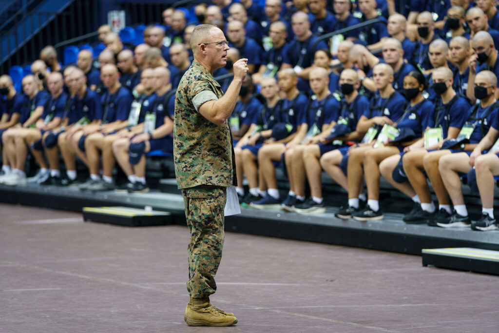 Military officer addressing a large group of seated cadets in an indoor arena.