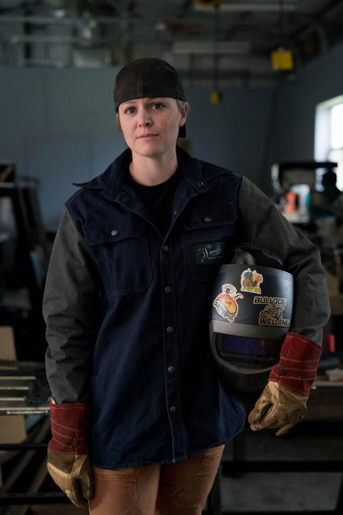 Woman in welding gear holds a welding helmet while standing inside a workshop.