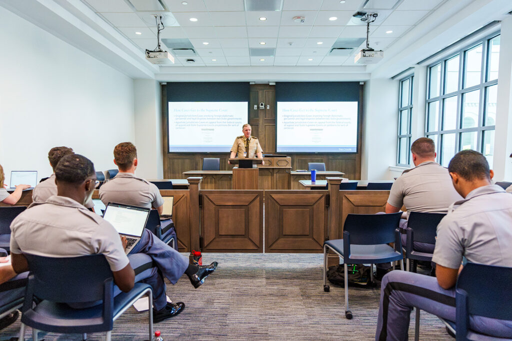 Instructor lectures to cadets in a courtroom-style classroom with presentation slides displayed on two screens.