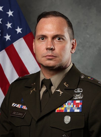 Dr. James Bezjian, in a U.S. Army dress uniform adorned with medals and ribbons, stands before an American flag, looking directly at the camera with a neutral expression.