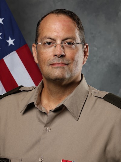 Dr. Robert J. Riggle, in a tan military-style uniform with glasses, stands before a U.S. flag, facing the camera with a neutral expression against a gray mottled background.