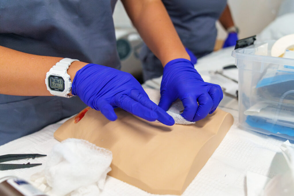 Nursing student wearing purple gloves practices wound care on a simulation model during a clinical skills lab.