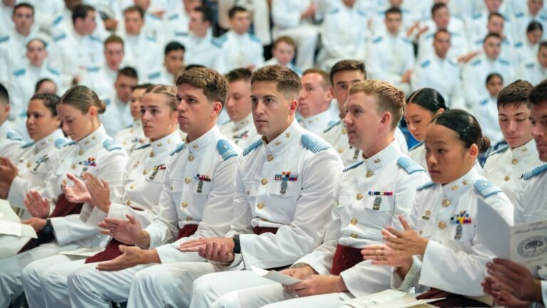 Cadets in dress white dress uniforms sit and applaud during an awards ceremony in McAlister Field House.