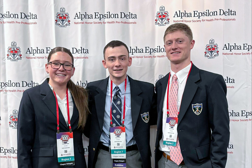 Cadets McKenna Magin, Dylan Horne and Banks Scarpa pose in front of an Alpha Epsilon Delta backdrop at the organization’s Centennial National Convention, wearing conference badges and Citadel blazers.
