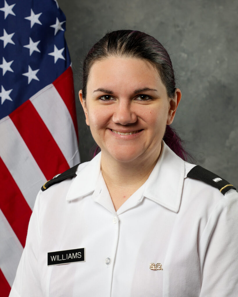 Portrait of Samantha Williams, lead instructor for maternal and newborn nursing in the Swain Department of Nursing, wearing a white Army duty uniform shirt with black shoulder boards. She stands in front of an American flag and gray studio backdrop, smiling at the camera. A name tag reading “WILLIAMS” and a small S.C. pin are visible on her uniform.