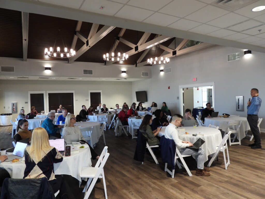 Faculty and staff sit at round tables working on laptops during a AI professional development workshop as a presenter, Jose Bowen, speaks at the front of the room.