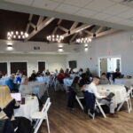 Faculty and staff sit at round tables working on laptops during a AI professional development workshop as a presenter, Jose Bowen, speaks at the front of the room.