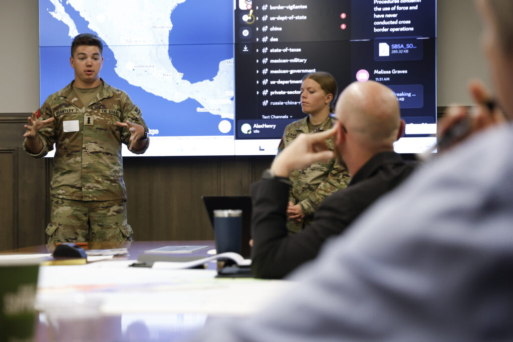 Two cadets stand in front of a digital map giving a presentation at a conference table during the Department of Intelligence and Security Studies Day of Intrigue.