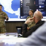 Two cadets stand in front of a digital map giving a presentation at a conference table during the Department of Intelligence and Security Studies Day of Intrigue.