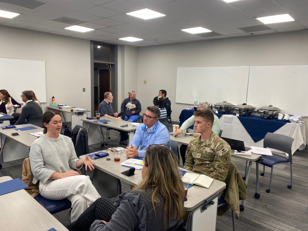A group of adults students sit at tables in a classroom, talking in small clusters.