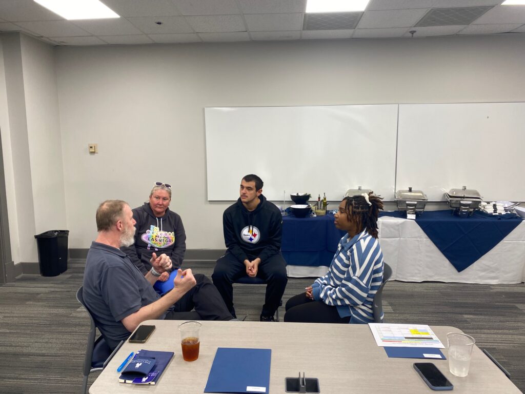 Four students sit in chairs in front of a table conferring.