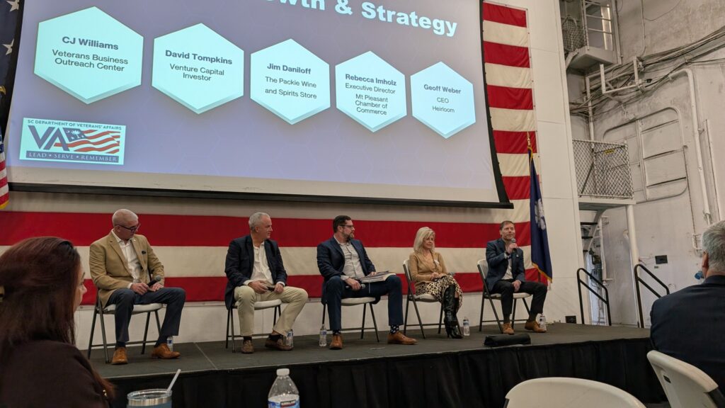Five panelists sit on a stage aboard the USS Yorktown during the South Carolina Department of Veterans’ Affairs Entrepreneurship Forum. A large screen behind them displays speaker names and affiliations, with an American flag hanging along the wall.