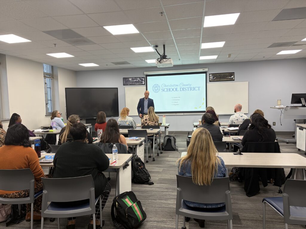 Assistant Education Professor Greg Harrison, Ph.D., stands at the front of a classroom, speaking to seated LIFT cohort participants during a session on school leadership, with a Charleston County School District presentation displayed on a screen behind him.