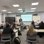 Assistant Education Professor Greg Harrison, Ph.D., stands at the front of a classroom, speaking to seated LIFT cohort participants during a session on school leadership, with a Charleston County School District presentation displayed on a screen behind him.
