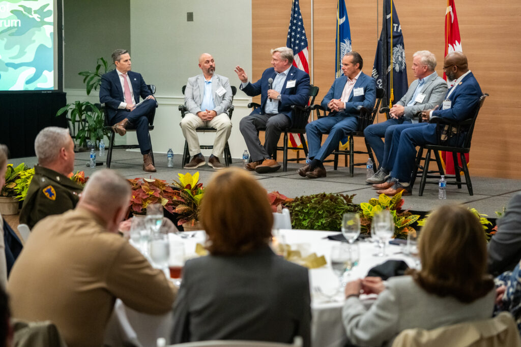 A panel of six guests in business attire sits on a stage at the Holliday Alumni Center during a Camo to Classroom discussion. American and state flags stand behind them as audience members seated at round tables watch from the foreground.