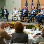 A panel of six guests in business attire sits on a stage at the Holliday Alumni Center during a Camo to Classroom discussion. American and state flags stand behind them as audience members seated at round tables watch from the foreground.