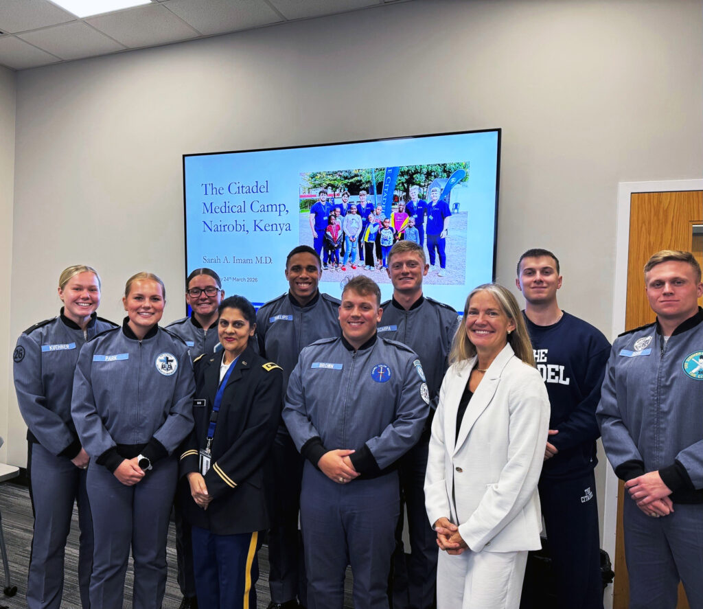 Retired Army Reserve Maj. Gen. Jami C. Shawley and Dr. Sarah Imam stand with cadets who participated in the Kenya Medical Camp, gathered in a classroom in front of a slide referencing the program in Kenya.