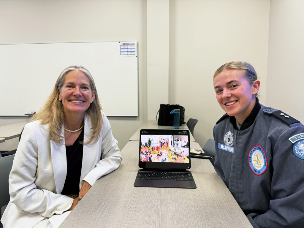 Retired Maj. Gen. Jami C. Shawley, U.S. Army Reserve, and 1st Battalion Commander Cadet Lt. Col. Lucy McArthur sit at a table during a one-on-one meeting, smiling toward the camera, with a tablet between them displaying images from McArthur’s Distinguished Scholars Project on women’s empowerment in Sub-Saharan Africa.