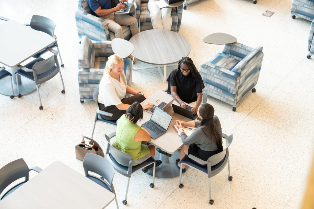 Four graduate students seated around a table collaborating on laptops in a campus study space.
