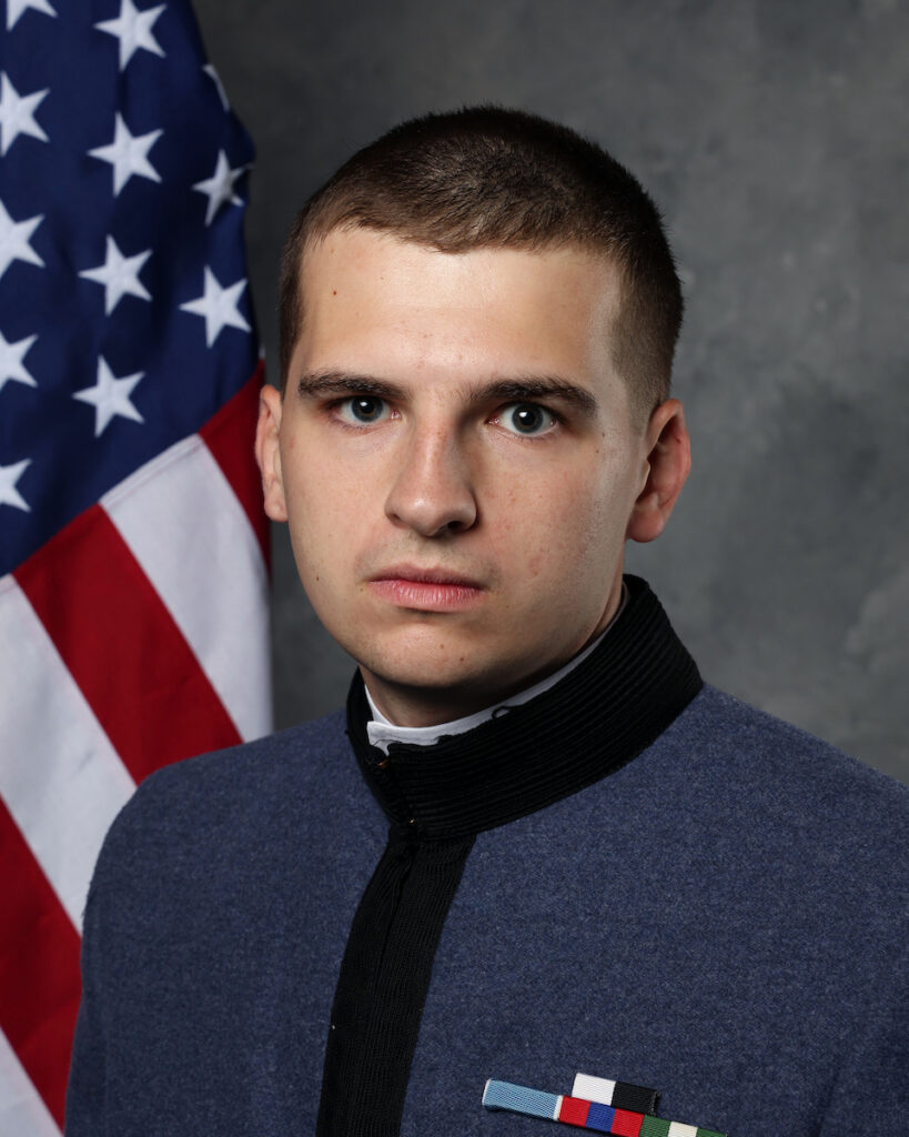 Formal portrait of a cadet in a gray military uniform with a high black collar, posed in front of a U.S. flag and gray studio backdrop.