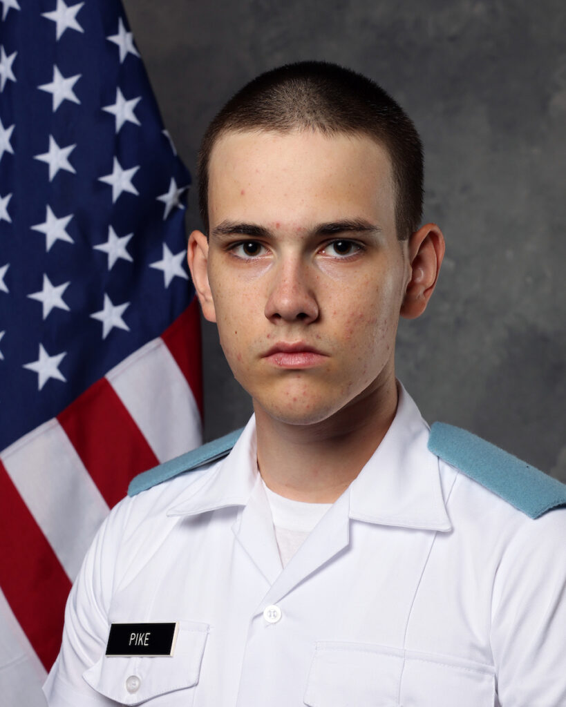Formal portrait of a cadet in a white short-sleeve military uniform with light blue shoulder boards, posed in front of a U.S. flag and gray studio backdrop.