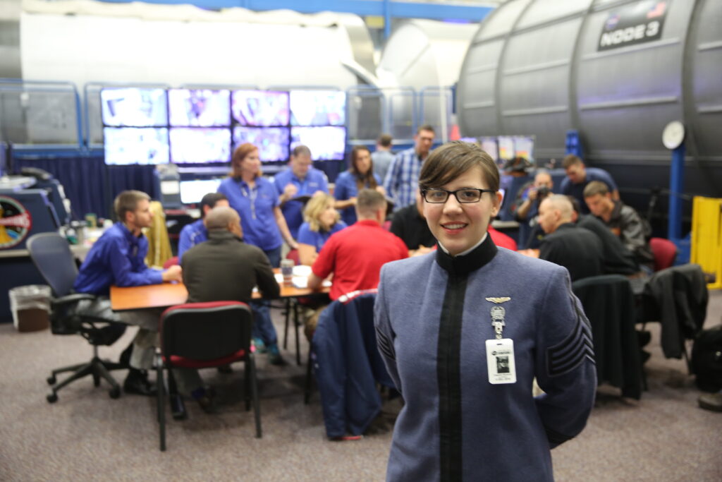 A Citadel cadet stands in a NASA training facility while a team works at tables and monitors in the background.