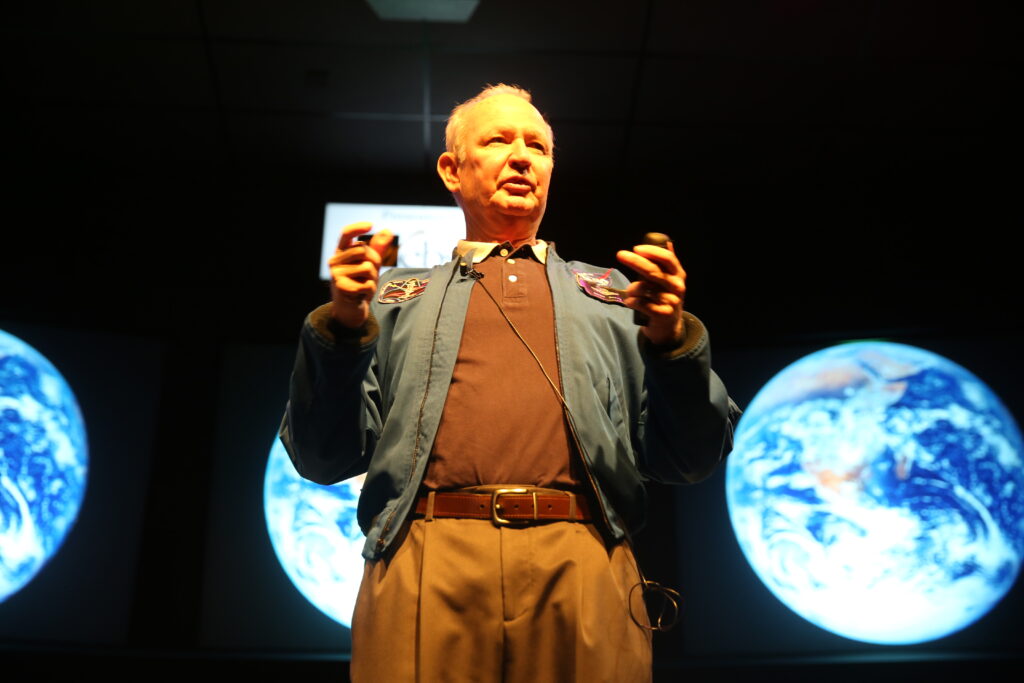 Astronaut Brian Duffy speaks during a presentation with images of Earth displayed on screens behind him.