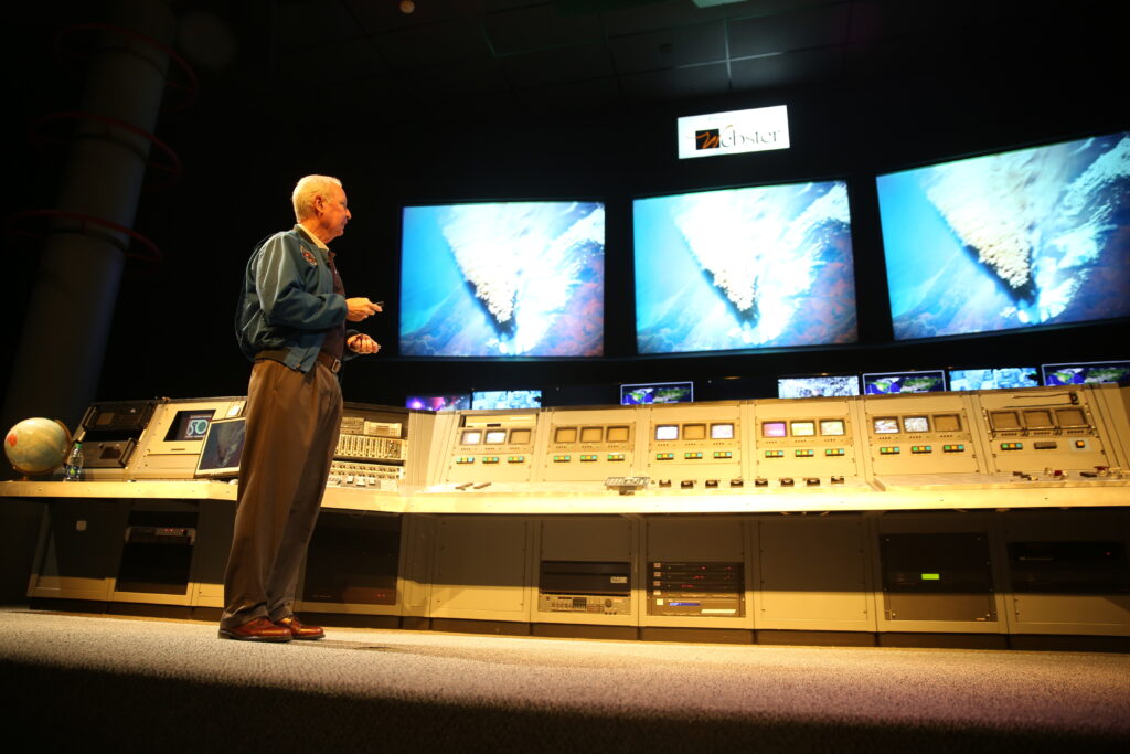 Astronaut Brian Duffy gives a presentation at Space Center Houston in front of multiple screens displaying Earth.