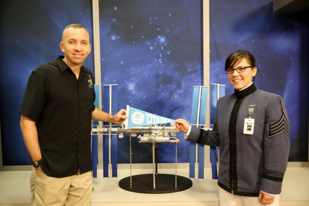 Randy Bresnik and a Citadel cadet hold a Citadel flag in front of a model of the International Space Station.