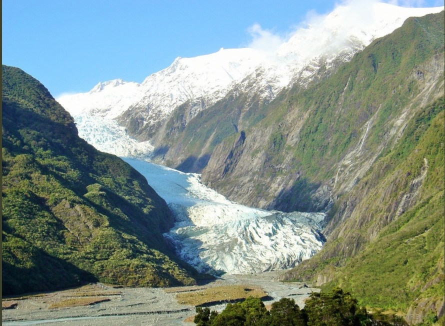 A glacier flows between steep green mountains beneath snow-covered peaks.