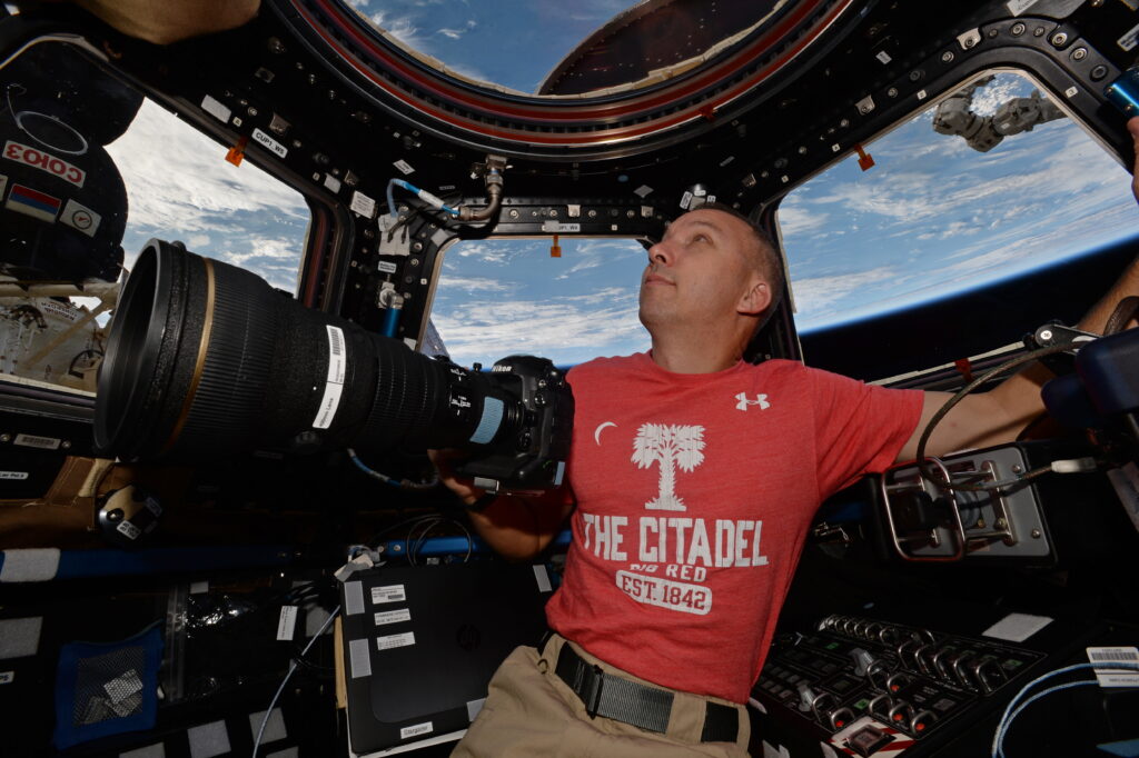Randy Bresnik holds a camera inside the International Space Station’s cupola while wearing a red Citadel T-shirt, with Earth visible through the windows.
