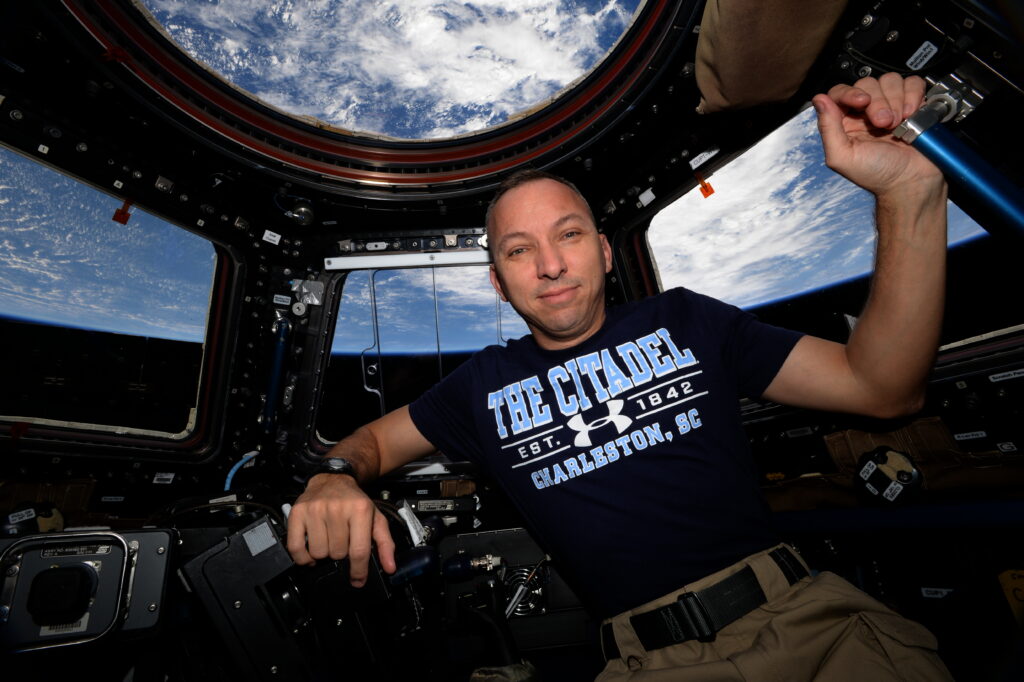 Randy Bresnik poses inside the International Space Station’s cupola wearing a navy Citadel T-shirt, with Earth visible through the windows.