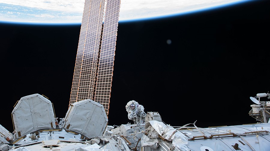 An astronaut performs a spacewalk outside the International Space Station, with a solar array and Earth visible in the background.