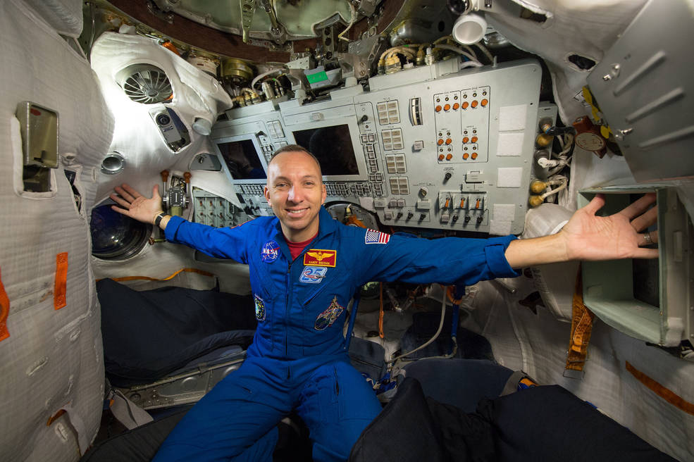 Randy Bresnik poses in a blue NASA flight suit inside a spacecraft surrounded by equipment and control panels.
