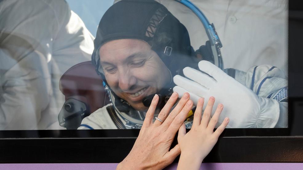 An astronaut inside a spacecraft smiles through a window while touching hands with family members on the other side of the glass.