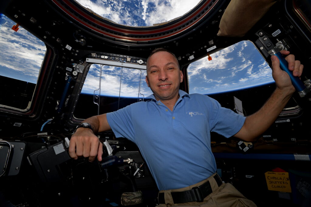 Randy Bresnik poses inside the International Space Station’s cupola wearing a light blue Citadel polo, with Earth visible behind him.