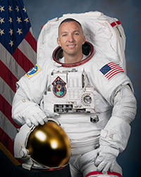 Astronaut in a white spacesuit poses for an official portrait with an American flag in the background.