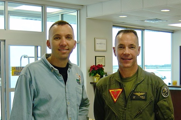 Two men stand together indoors, one wearing a military flight suit.