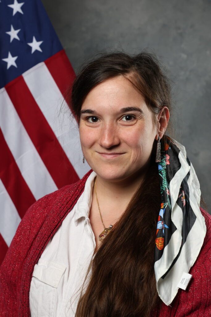 A person with long brown hair smiles while wearing a red sweater and a patterned scarf, standing in front of an American flag.
