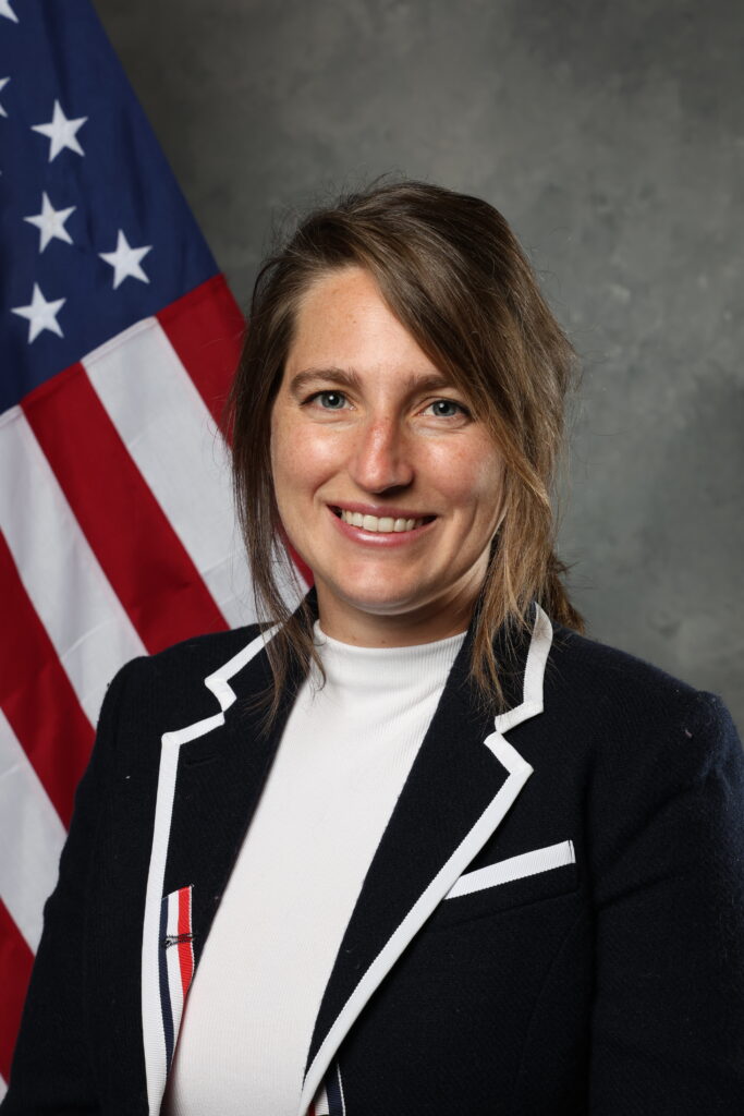 A smiling person in a dark blazer with white trim stands in front of an American flag.