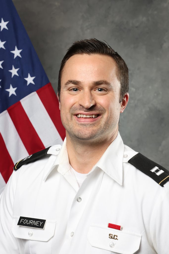 A person in a white uniform with name tag "FOURNEY" and insignia stands before an American flag.